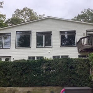 A daytime, eye-level, medium shot captures the rear exterior of a two-story, white-sided house nestled among lush green trees under an overcast sky